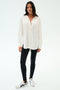 A woman stands against a white background wearing the SPLITS59 Kate Airweight Lite Button Down in white, styled as a chic layering piece with black leggings and white sneakers featuring black stripes.