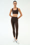 A person stands facing forward in the SPLITS59 Lexi Rigor Bra - Dark Chocolate/Black with matching leggings and white sneakers—ideal gym wear—against a plain white background.
