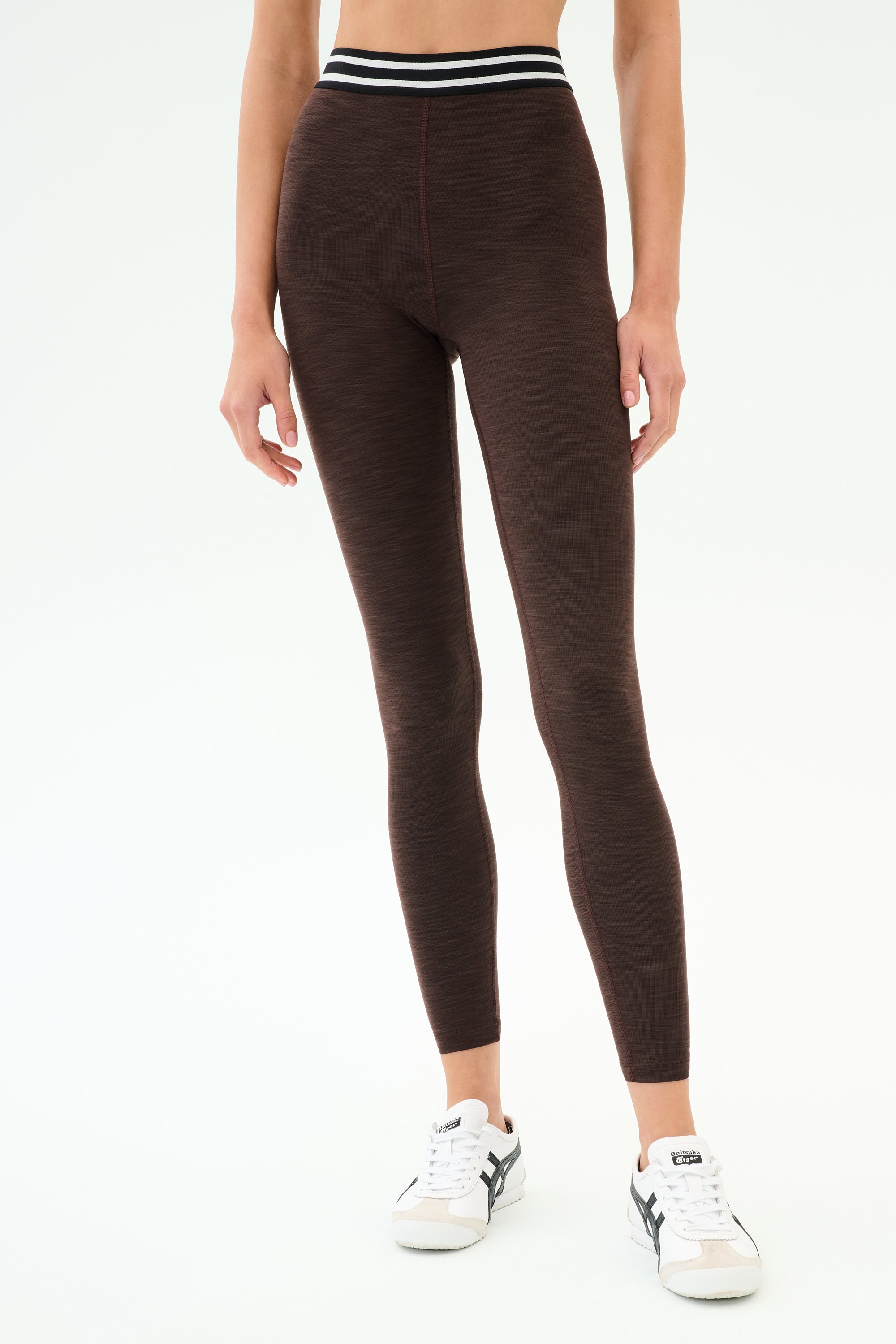 A person wearing SPLITS59 Bailey High Waist Active Rib 7/8 leggings in Heather Dark Chocolate with a striped waistband and white sneakers stands against a plain white background.