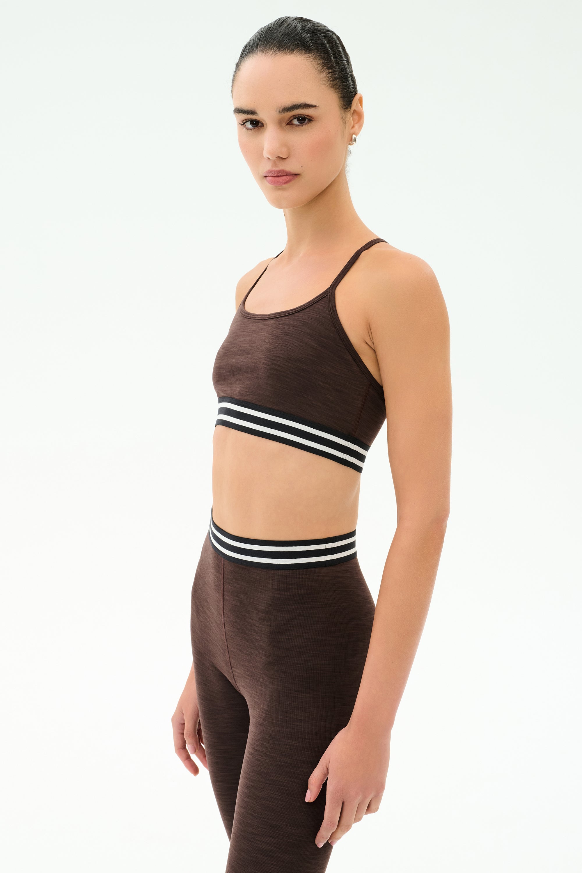A woman wears the SPLITS59 Cali Active Rib Bra in Heather Dark Chocolate with matching leggings featuring black and white striped bands, standing against a plain white background.