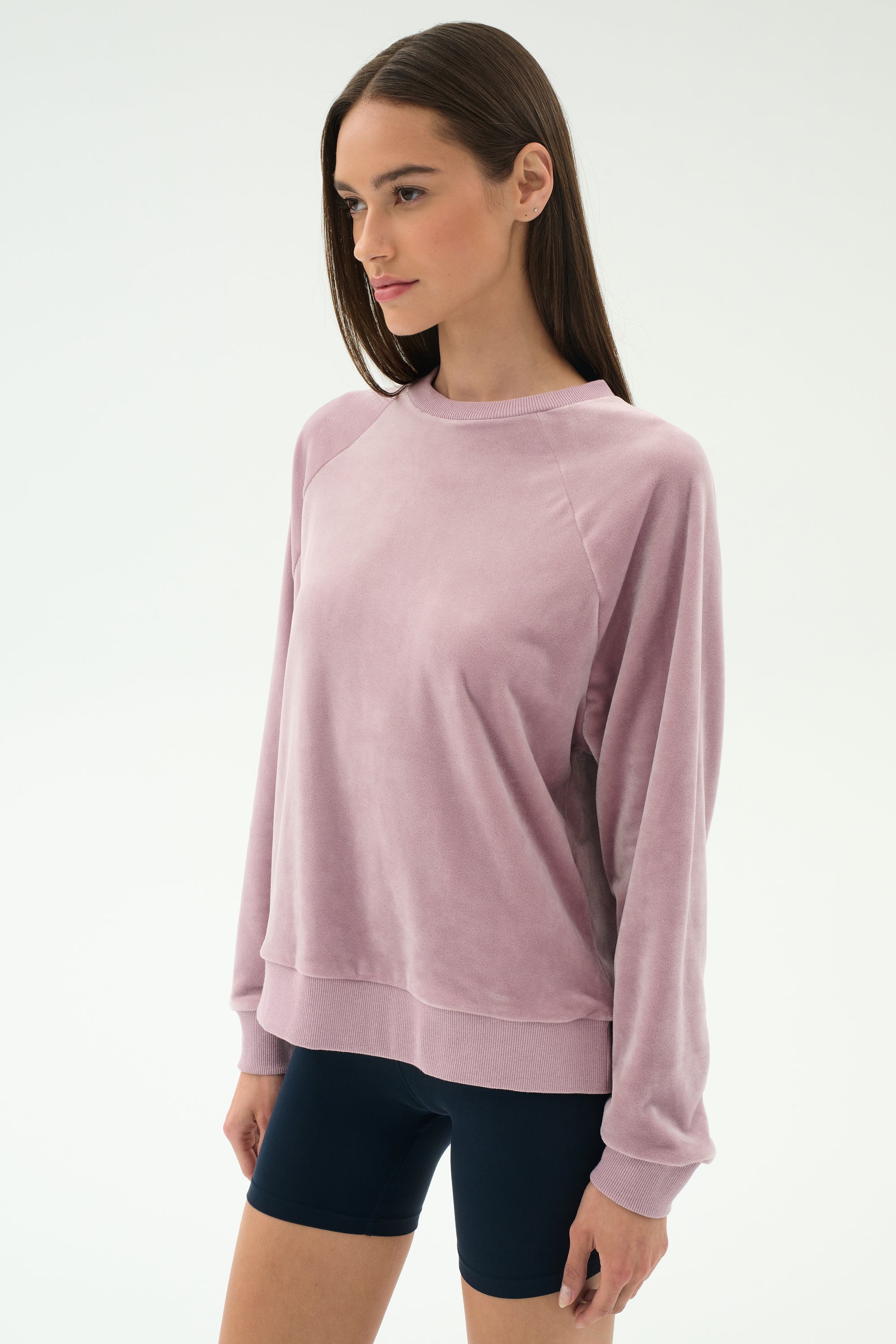 A woman wears the SPLITS59 Joy Velour Crewneck in Blush and black spandex shorts, standing against a plain white background.