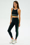 A person in the SPLITS59 Layla Airweight High Waist 7/8 athletic outfit in Black/Arugula, made from breathable spandex, stands against a plain background, looking to the side while wearing white sneakers.