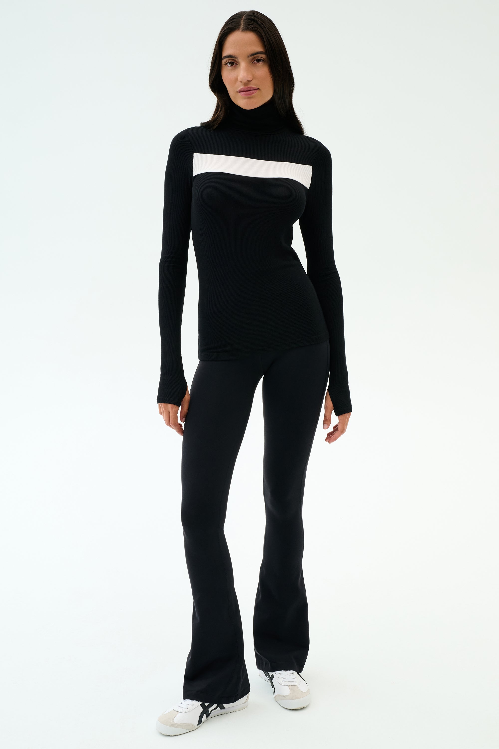 A woman faces forward wearing the SPLITS59 Lennon Rib Turtleneck in Black/White with black flared yoga pants and white sneakers against a plain light background.