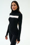 A woman wears the SPLITS59 Lennon Rib Turtleneck in Black/White, featuring a white horizontal chest stripe, standing against a plain light background—perfect for yoga or running.