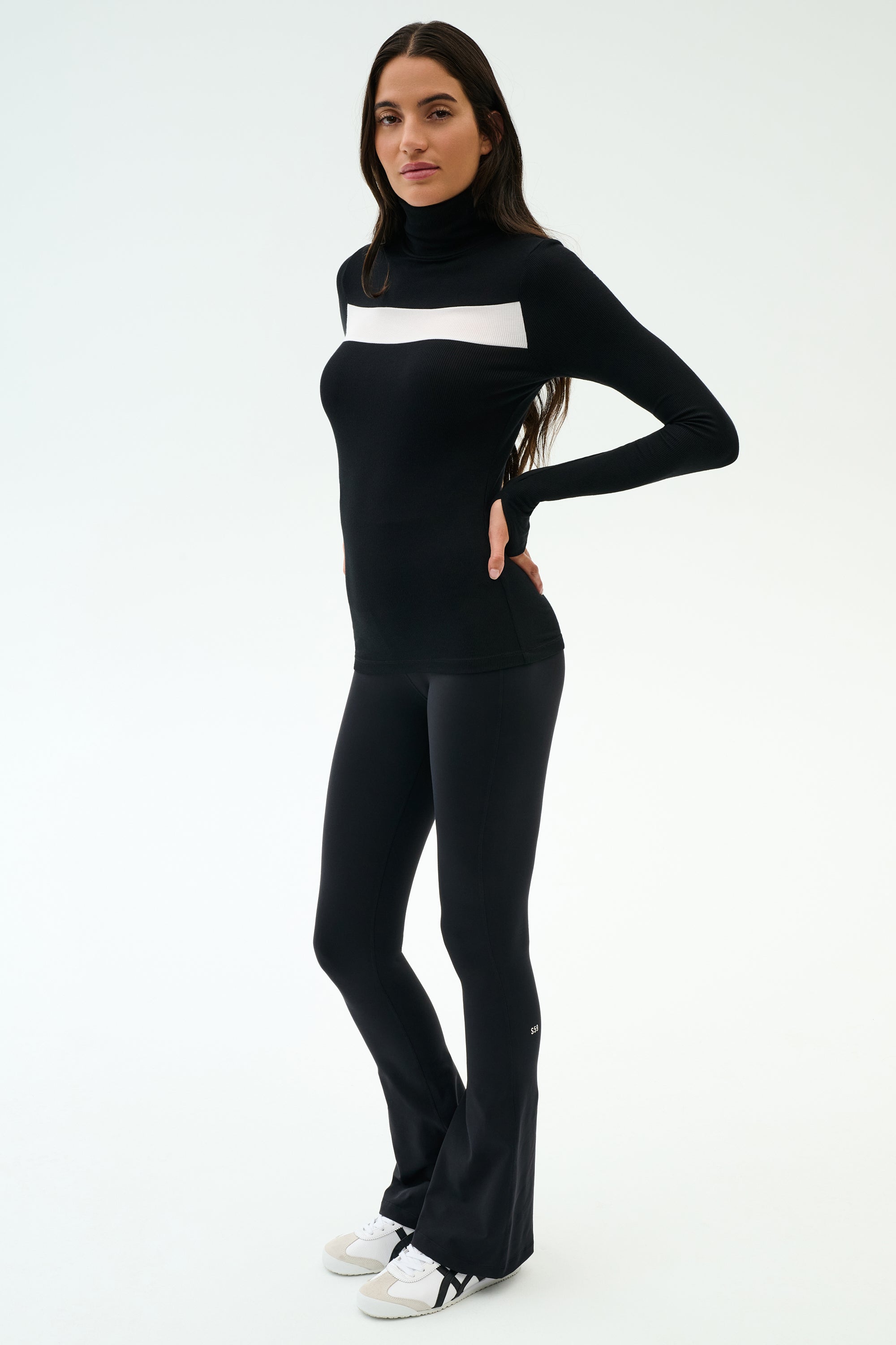 A woman poses against a plain background in the SPLITS59 Lennon Rib Turtleneck - Black/White, pairing it with black flared pants and white sneakers for a effortlessly stylish, yoga-inspired look.