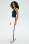 A person with curly hair in a navy tank top and SPLITS59's Maxie Rigor Track Pant in White/Indigo, featuring a dark stripe, stands looking slightly sideways in a studio setting.