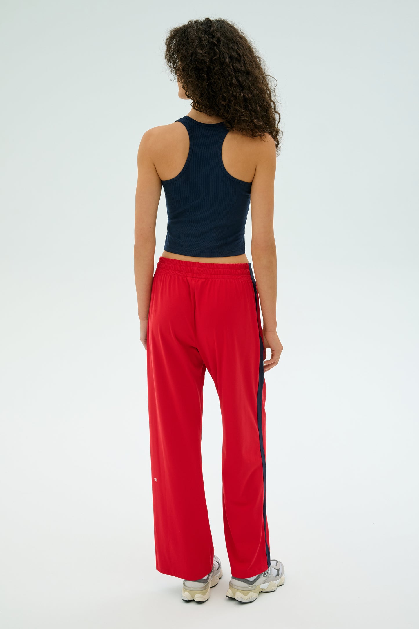 With curly hair, a person dons a black tank top and vibrant Pirate Red/Indigo Maxie Rigor Track Pant by SPLITS59. They pair it with white sneakers, standing back to the camera against a plain backdrop for an effortlessly casual vibe.