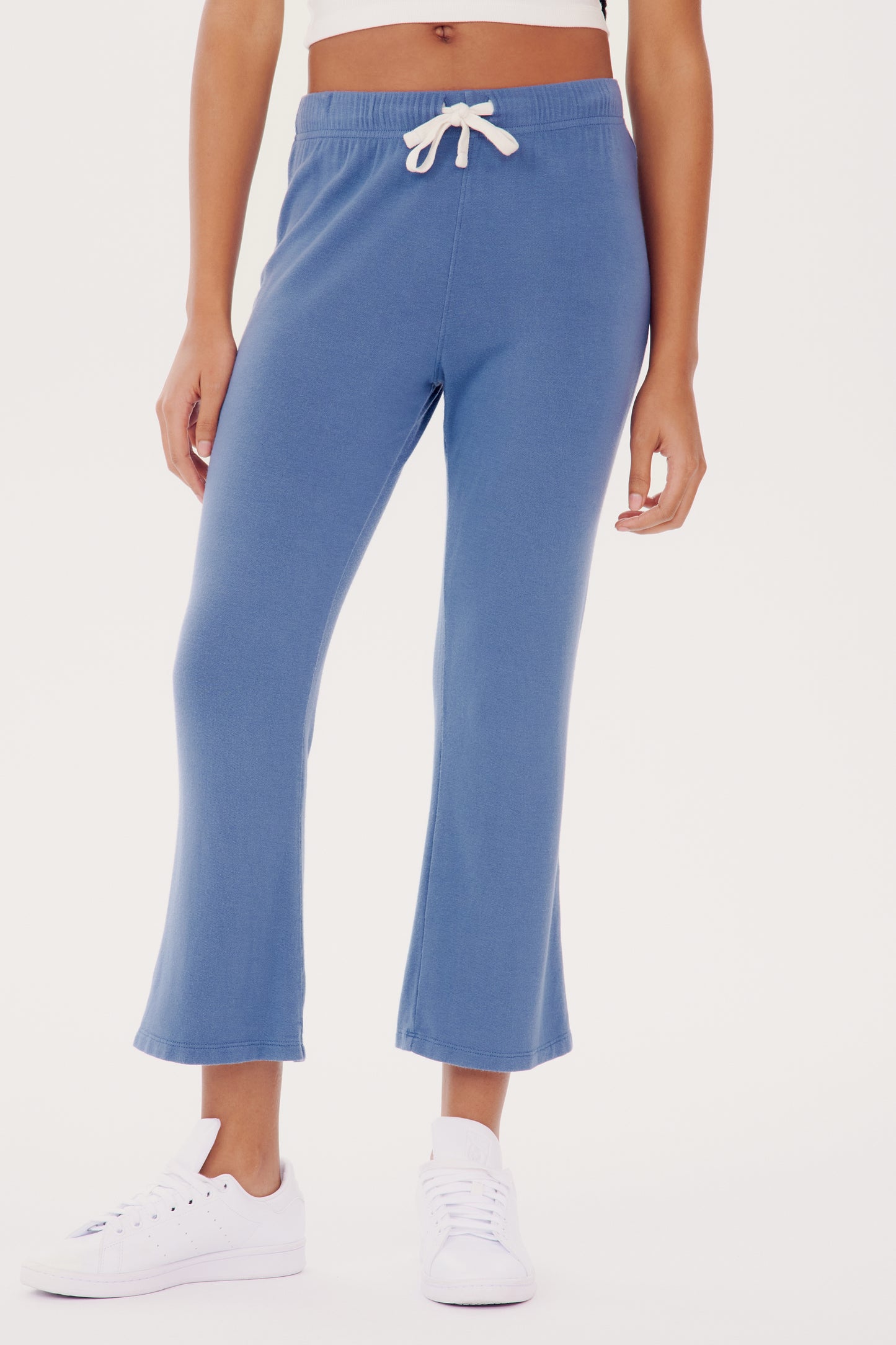 Woman standing in SPLITS59 Brooks Fleece Cropped Flare - Steel Blue sweatpants and white sneakers, cropped view showing only from waist to shoes.