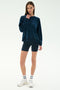 A woman stands against a plain background wearing the SPLITS59 Joy Velour Crewneck in Indigo, navy cycling shorts, white socks, and white athletic shoes.