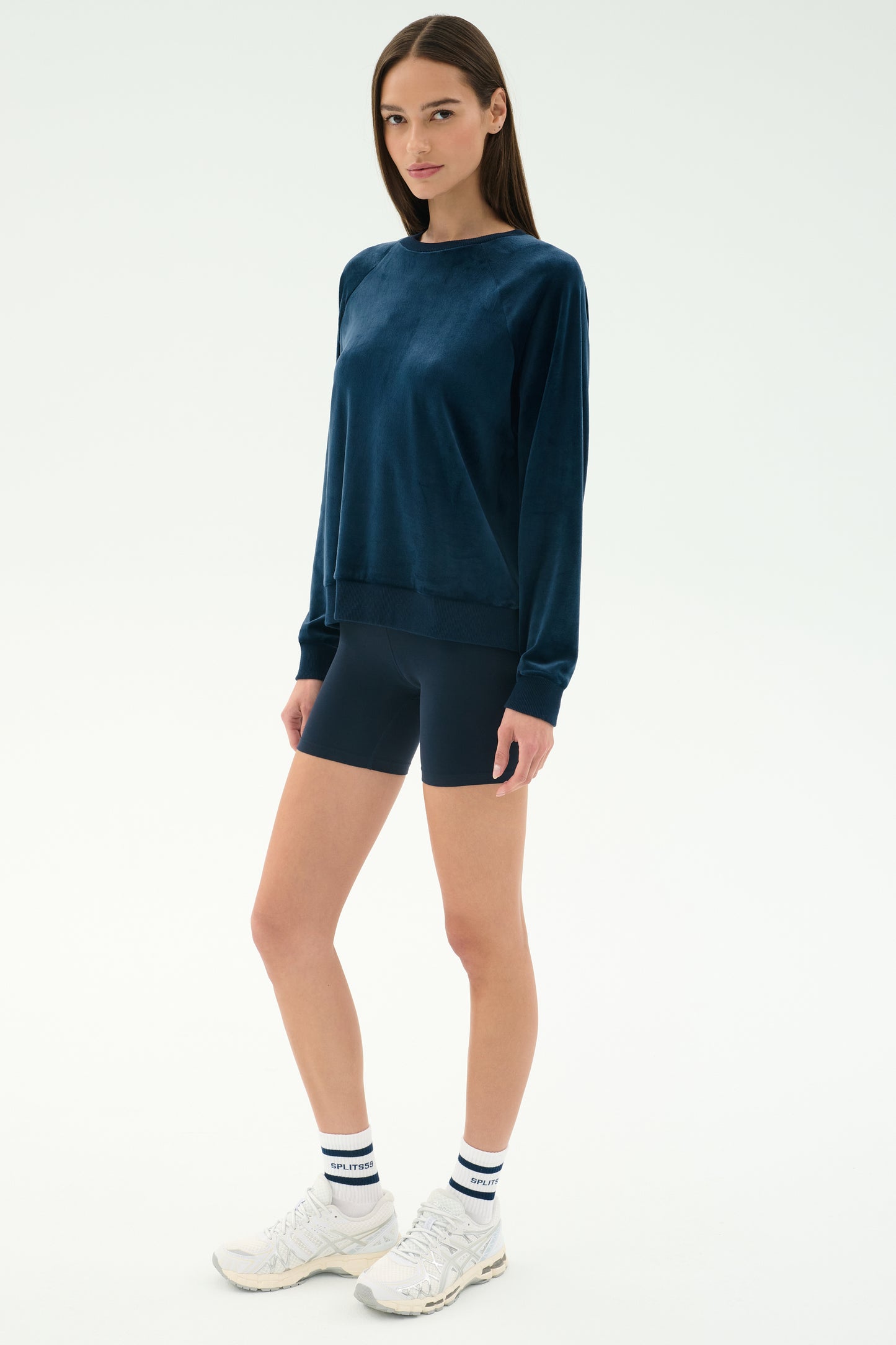 A woman stands against a plain background wearing the SPLITS59 Joy Velour Crewneck in Indigo, black cycling shorts, white socks, and white sneakers.