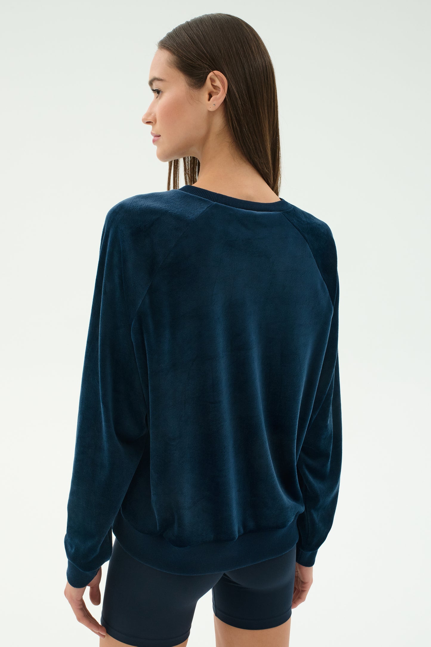 A woman with straight brown hair is seen from behind wearing the SPLITS59 Joy Velour Crewneck in Indigo and black shorts against a plain light background.