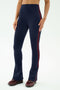 A person models SPLITS59's Raquel High Waist Flared Legging in Indigo/Dark Cherry, featuring a flared silhouette and side stripe, paired with burgundy sneakers against a plain background.