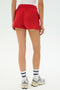 Model wearing SPLITS59 Reena Sweatshort in Pirate Red, white top, white sneakers, and white socks with blue stripes, standing on a white background, shown from the back.