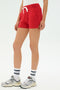 Model wears the SPLITS59 Reena Sweatshort in Pirate Red with white crew socks featuring navy stripes, gray athletic sneakers, and a white top, posed against a plain white background.