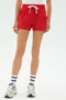 Model wears the SPLITS59 Reena Sweatshort in Pirate Red, paired with white sneakers, blue-striped white athletic socks, and a white top, standing against a plain light background.