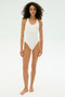 A woman with curly hair stands barefoot against a plain white background, wearing the SPLITS59 Suki Airweight Bodysuit in white, a smooth nylon sleeveless one-piece.