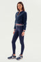 A person stands in a full-body shot, wearing a cozy SPLITS59 Willa Sherpa Jacket - Indigo with a striped collar, dark leggings, and sneakers, against a plain white background. They look ready for winter workouts as they warm up for their routine.