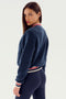 A woman with straight hair wearing a navy blue fleece jacket with red, white, and blue striped trim stands facing slightly to the side, looking at the camera, showcasing the Willa Sherpa Jacket - Indigo by SPLITS59, perfect for winter workouts or supporting her favorite sports team.