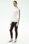 A woman stands against a plain background wearing the SPLITS59 Jackson Rib Full Length Turtleneck in White/Dark Chocolate, brown leggings, and white athletic shoes—ideal for stylish yoga or running sessions.