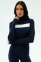 A woman stands against a plain light background wearing the SPLITS59 Lennon Rib Turtleneck in Indigo/White, featuring a navy blue ribbed design with a white horizontal stripe across the chest.