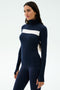 A woman stands against a plain light background wearing the SPLITS59 Lennon Rib Turtleneck in Indigo/White, featuring a fitted silhouette and white horizontal chest stripe, paired with matching navy leggings.
