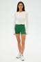 A woman is wearing the Ella Airweight High Waist Short 3.5" in Arugula/White by SPLITS59 with a white long-sleeve top tied at the waist and white sneakers, standing against a plain background with relaxed arms.