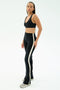 A person wearing the SPLITS59 Raquel High Waist Flared Legging in Black/Creme and a black sports bra with white sneakers stands against a plain white background.