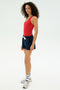 A woman in a red tank top, striped socks, and sneakers wears SPLITS59's Reena Sweatshort in Indigo, standing against a plain white background.