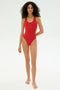 A woman stands barefoot against a plain background, wearing the SPLITS59 Suki Airweight Bodysuit in Pirate Red.