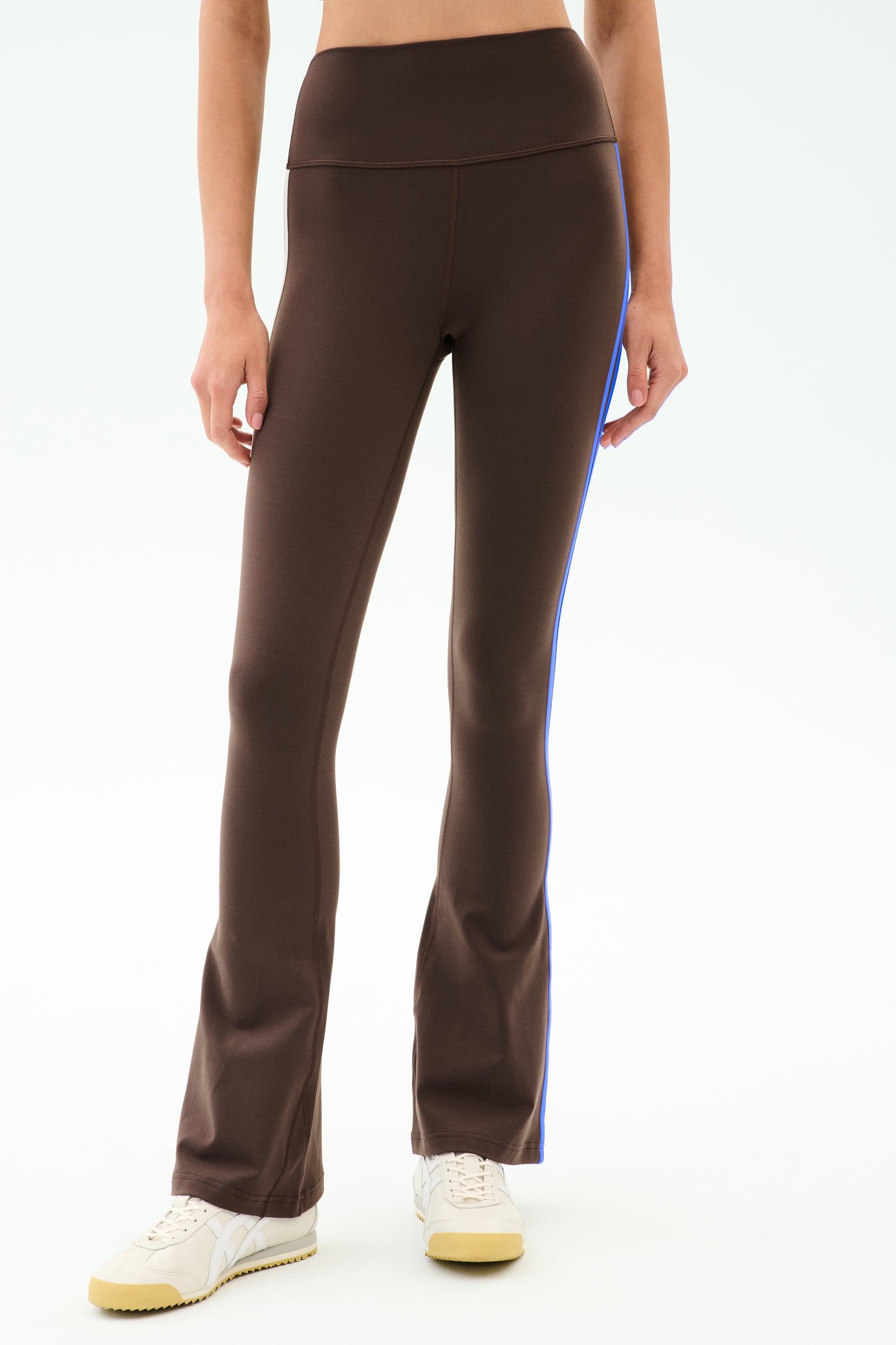A model is shown from waist to feet wearing SPLITS59 Raquel High Waist Supplex Flare in Dark Chocolate/Classic Blue, paired with white sneakers and a crop top. The 4-way stretch supplex fabric ensures comfort for workouts or all-day wear.