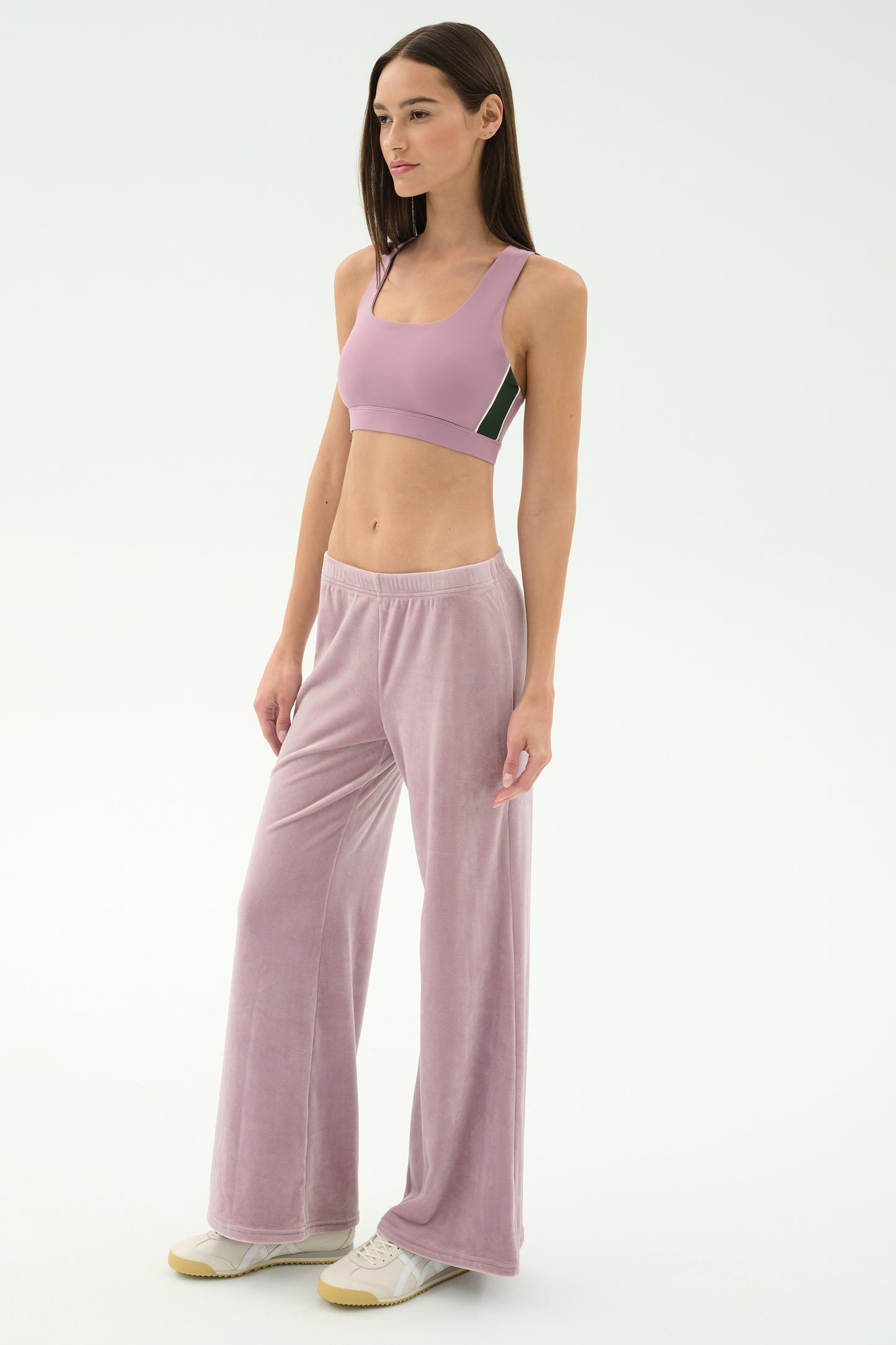 A woman stands in SPLITS59’s Joy Velour Pant in Blush, paired with a light pink sports bra and white sneakers, against a plain white background.