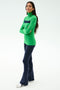 A woman stands with arms crossed, wearing the SPLITS59 Lennon Rib Turtleneck in Grass/Indigo, navy flared pants, and white sneakers against a plain light background.