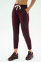 Someone wearing the SPLITS59 Reena Fleece 7/8 Sweatpant in Dark Cherry, featuring soft fleece fabric and white drawstrings, paired with matching burgundy sneakers, stands hands-in-pockets against a plain background.