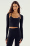 A woman with long brown hair is wearing the SPLITS59 Shiri Airweight Shrug - Black, a stylish long-sleeve crop top with thumbholes, paired with black high-waisted leggings. Both pieces are crafted from a comfortable blend of nylon and spandex. She stands facing forward against a plain white background.