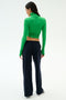 A woman stands facing away in a green long-sleeve turtleneck crop top, SPLITS59 Brynn Airweight Pant in Indigo, and white sneakers against a plain white background.
