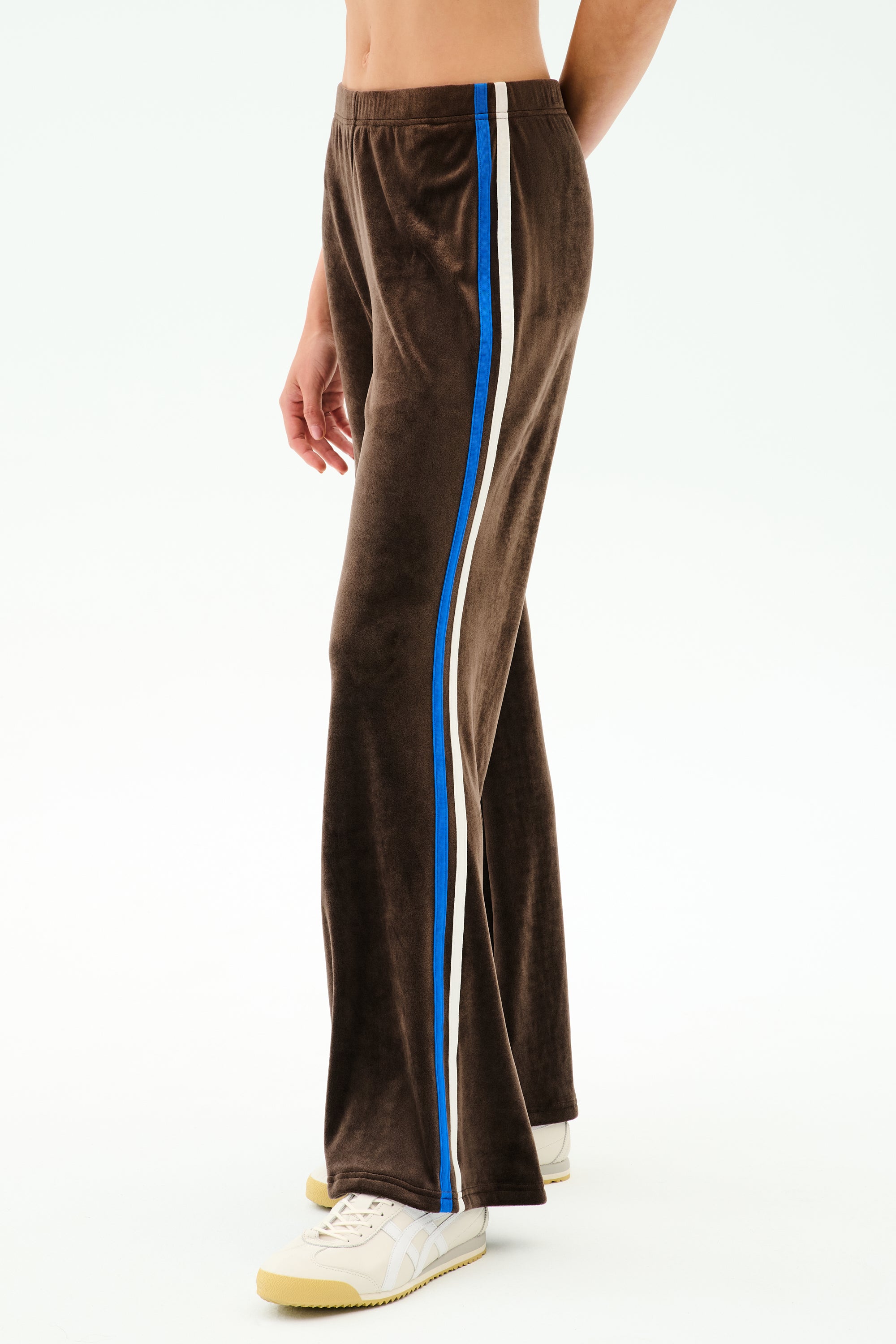 A person wears the SPLITS59 Heritage Velour Pant in Dark Chocolate/Classic Blue with blue and white side stripes, paired with white sneakers and a bare midriff, standing against a plain white background.