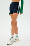 Wearing a green long-sleeve top, SPLITS59 Ella Airweight High Waist Short in Indigo/White, white socks, and white sneakers.