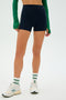 A person in a green long-sleeve top wears SPLITS59's Ella Airweight High Waist Short 3.5" in Indigo/White, standing with legs apart in white sneakers and green-striped socks against a plain white background.