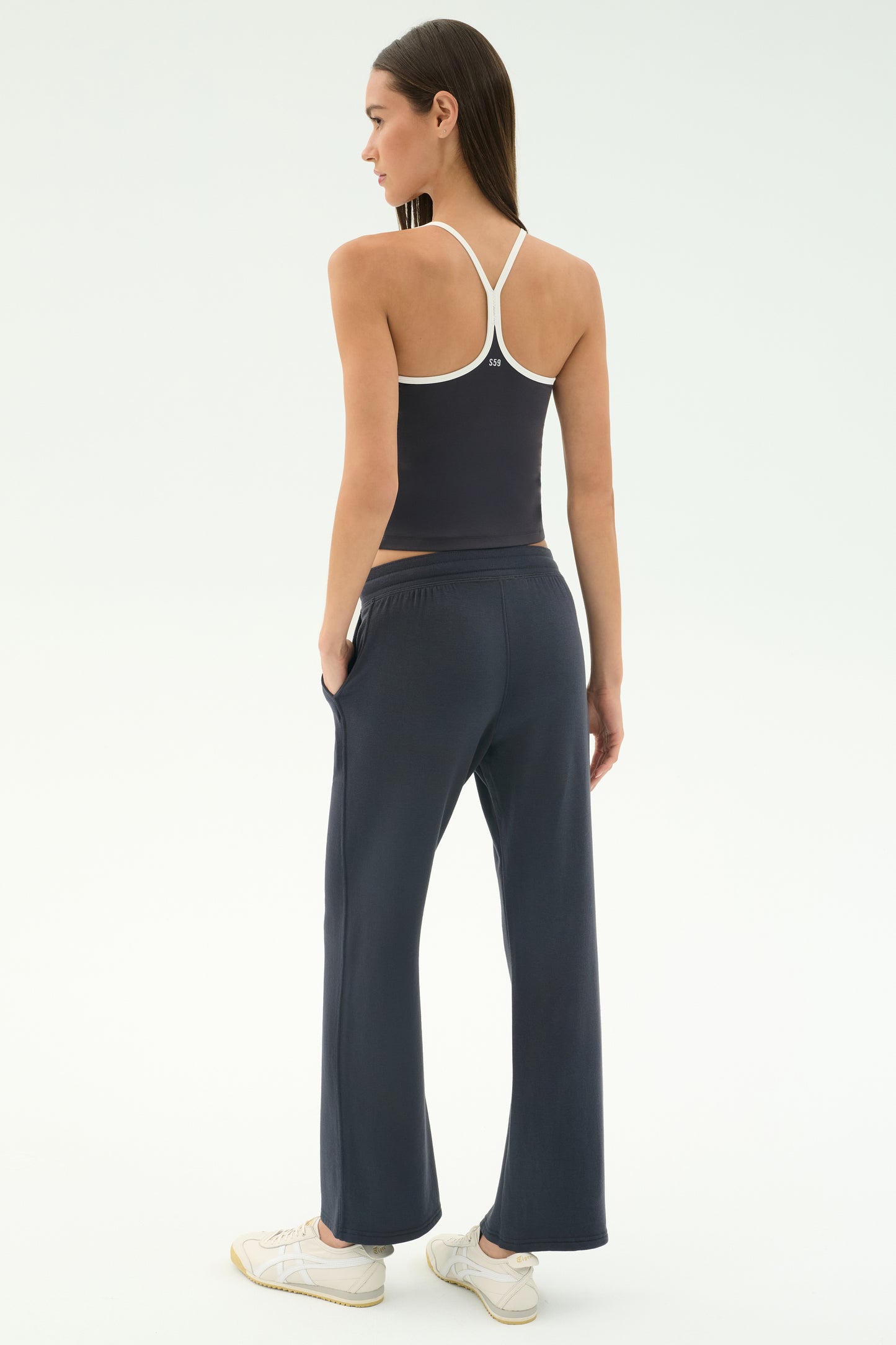 A woman faces away, wearing a navy racerback tank top and SPLITS59 Raven Fleece Full Length Sweatpant in Graphite with white sneakers against a plain light background.