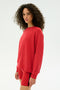A woman with curly hair wears the SPLITS59 Andie Fleece Sweatshirt in Pirate Red, paired with matching red shorts featuring a black stripe, standing against a plain white background.