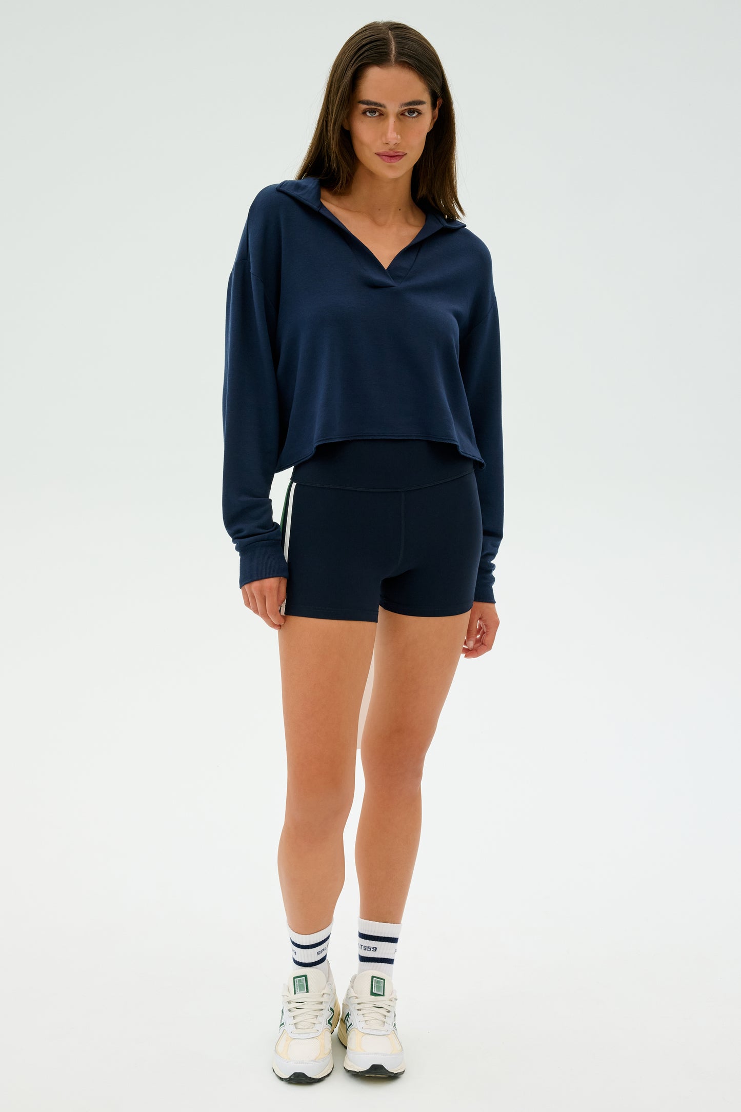 A woman in a navy Joan Fleece Polo Sweatshirt - Indigo by SPLITS59 and black shorts stands against a plain background, wearing white sneakers and socks.