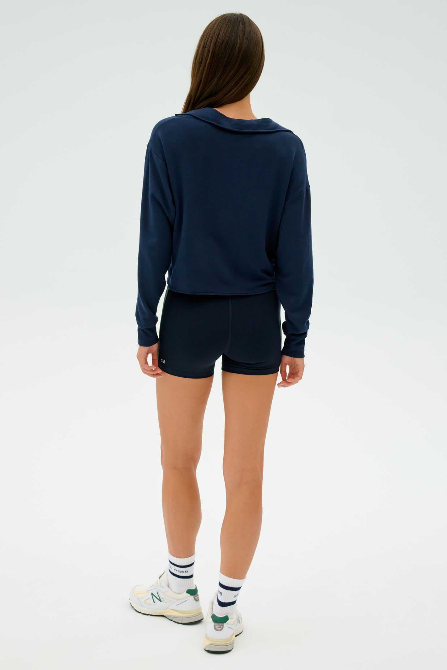 A woman with long brown hair stands on a plain white background, wearing the SPLITS59 Joan Fleece Polo Sweatshirt in indigo and soft modal shorts. She completes her look with white sneakers and striped socks, facing away from the viewer.
