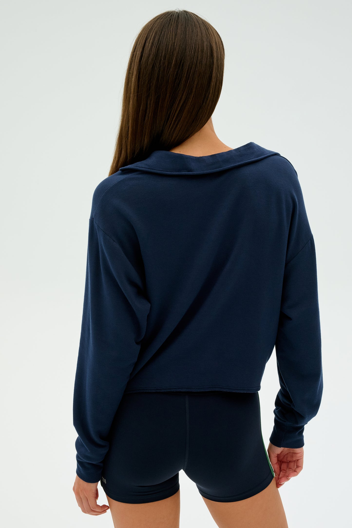 A person with long hair in an indigo Joan Fleece Polo Sweatshirt by SPLITS59 and black shorts stands against a plain background, facing away.