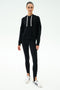 A person stands facing forward wearing the SPLITS59 Joey Long Sleeve Hoodie in black, paired with black spandex-blend stirrup leggings and white sneakers with black details, set against a plain white background.