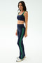 A woman stands in a studio wearing SPLITS59’s Raquel High Waist Flared Legging in Indigo/Grass, featuring green stripes and 4-way stretch fabric for ultimate comfort—perfect for any workout. She pairs them with a matching sports bra and white sneakers.
