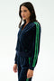 A woman models the SPLITS59 Heritage Velour Bomber in Indigo/Grass, featuring green stripes on navy blue velour, combining sporty comfort with yoga-inspired style against a white background.