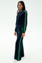 Woman in the SPLITS59 Heritage Velour Bomber - Indigo/Grass, featuring navy velour with green stripes on sleeves and legs, perfect for yoga or lounging, poses against a white background.
