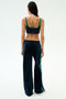 A woman stands facing away against a plain light background, wearing the SPLITS59 Heritage Velour Pant in Indigo/Grass—navy wide-leg pants with green side stripes that skim her ankles.