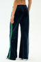 A person is seen from behind wearing the SPLITS59 Heritage Velour Pant in Indigo/Grass, featuring navy velour fabric with green side stripes and a relaxed wide-leg fit that flares stylishly at the ankle, against a plain white background.