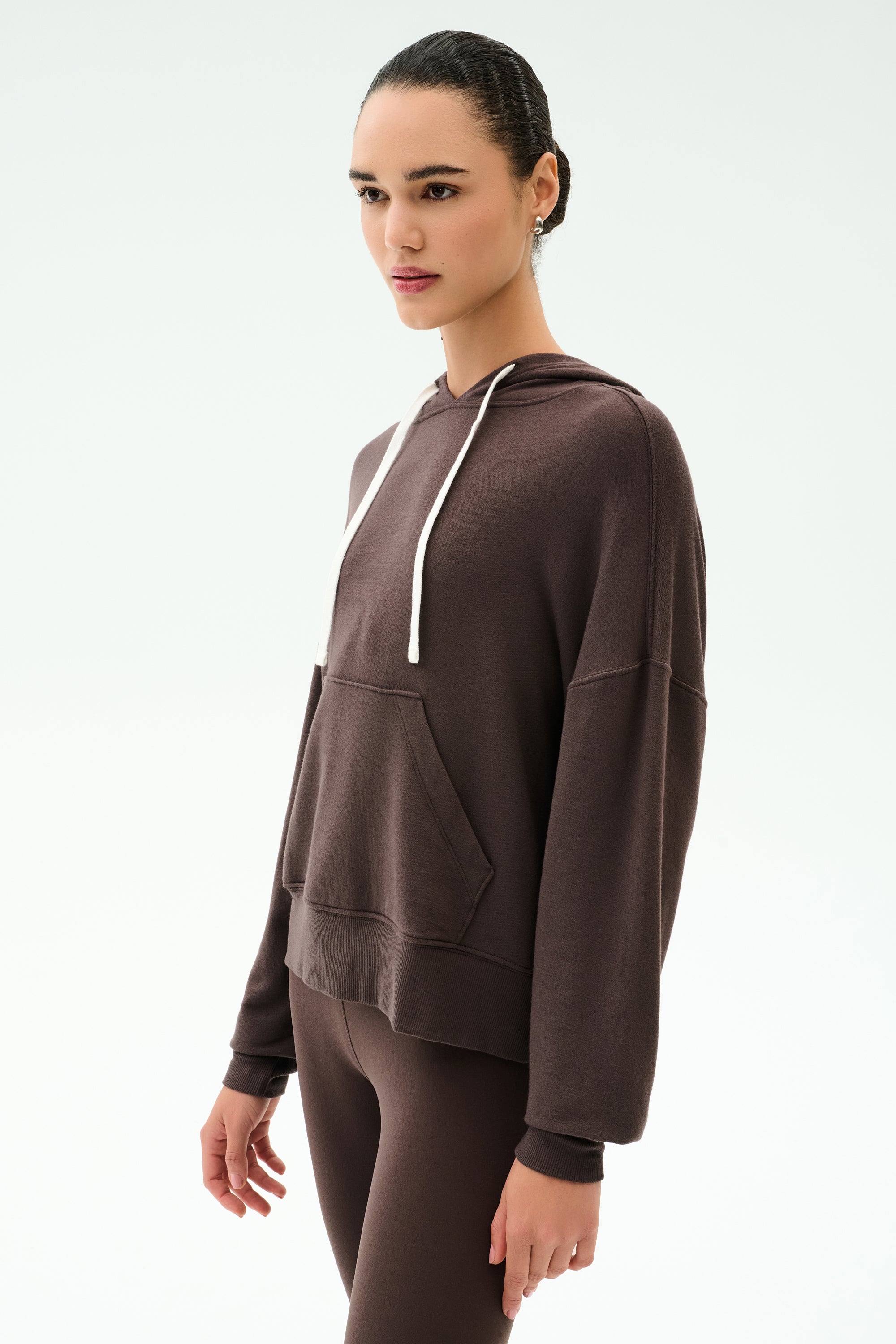 A woman wears the SPLITS59 Joey Long Sleeve Hoodie in Dark Chocolate with matching brown leggings, both crafted from a soft modal spandex blend, standing against a plain light background.